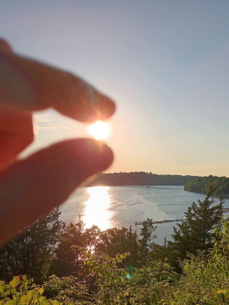 Sun being held between the thumb and forefinger with a forest and lake in the background