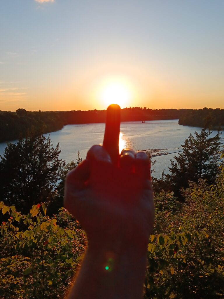 Sun on the end of a middle finger with a forest and lake in the background